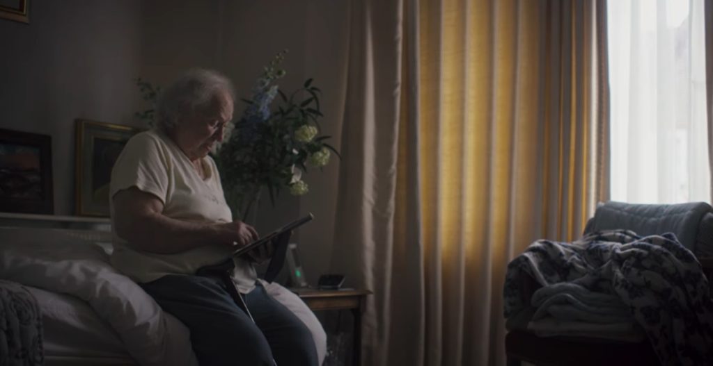 An elderly woman sits on bed and looks at an image.